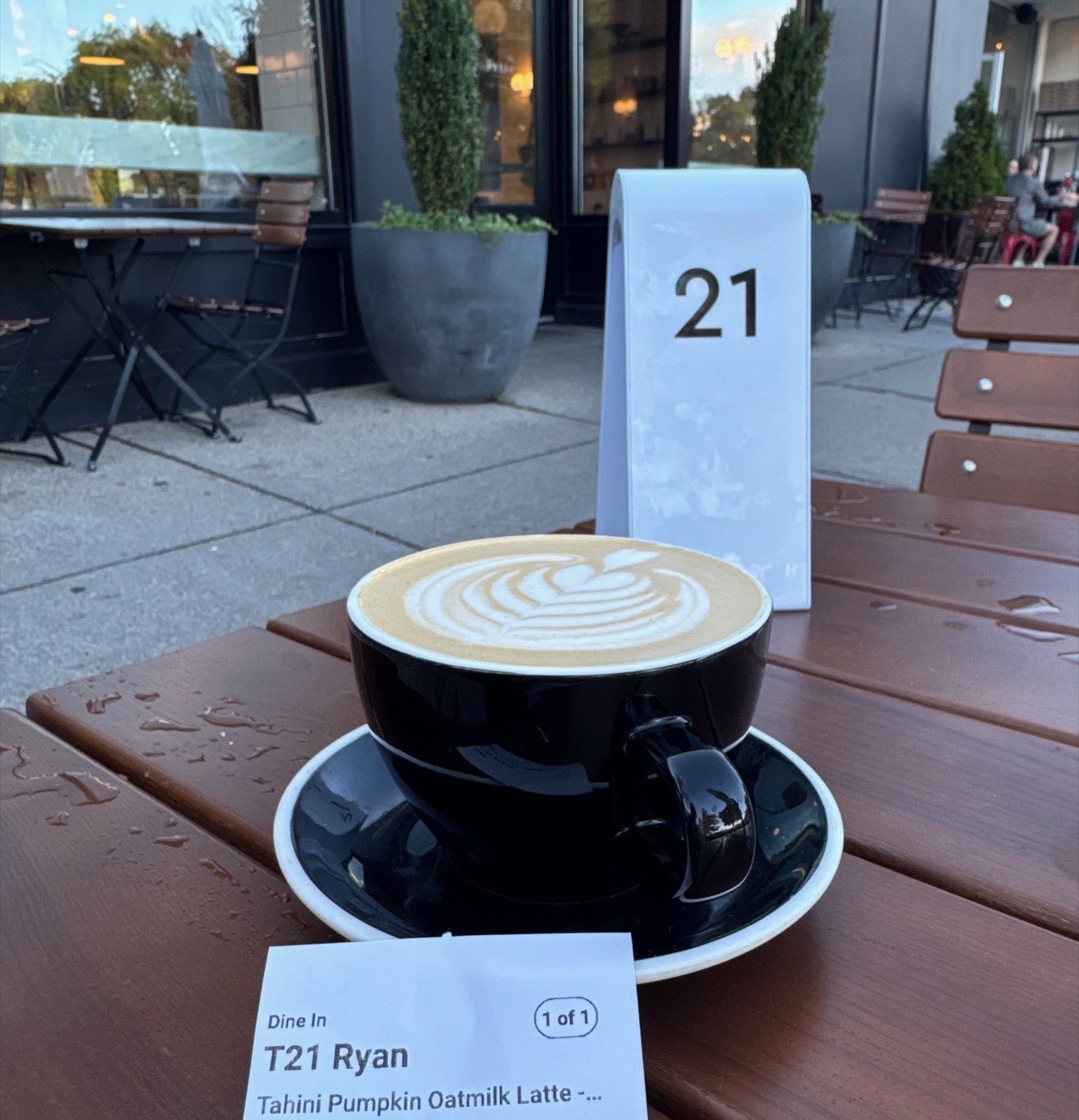 A Tahini Pumpkin Oatmilk Latte at Tatte: a black cup on a saucer with rosetta latte art, on a wooden patio table, with the order ticket naming the drink in the foreground.