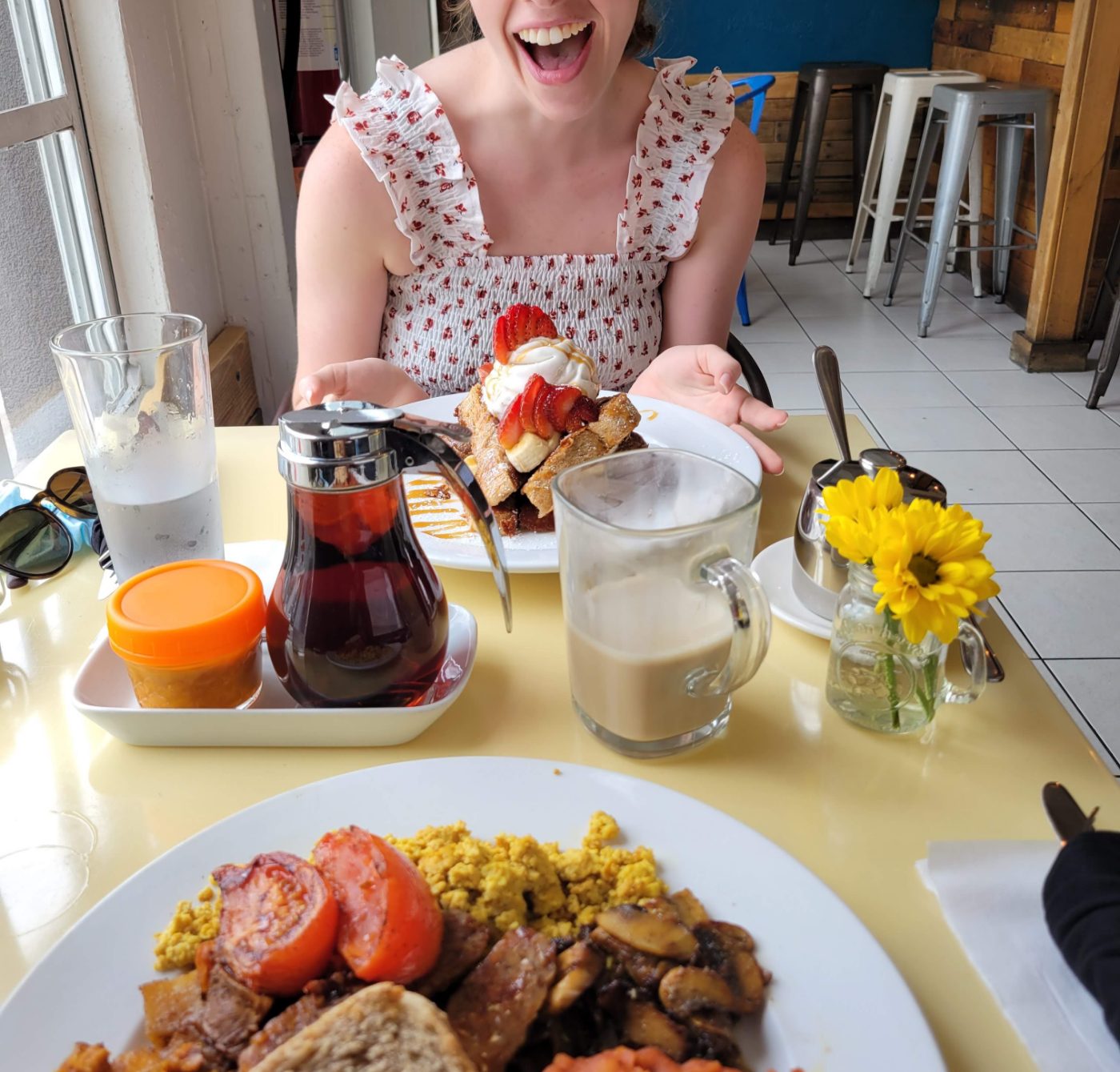 A vegan brunch for two on a yellow diner table: French toast stacked with strawberries, banana and whipped cream, a jar of maple syrup, a milky latte in a glass mug, a small vase of yellow flowers, and in the foreground a plate of tofu scramble, roasted tomatoes, mushrooms and toast.